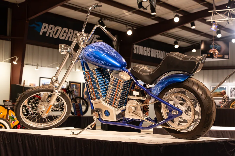 The Shandoka electric chopper on the Sturgis Buffalo Chip stage, side on, its long blue frame carrying the stacked battery modules where a V-twin engine would sit.