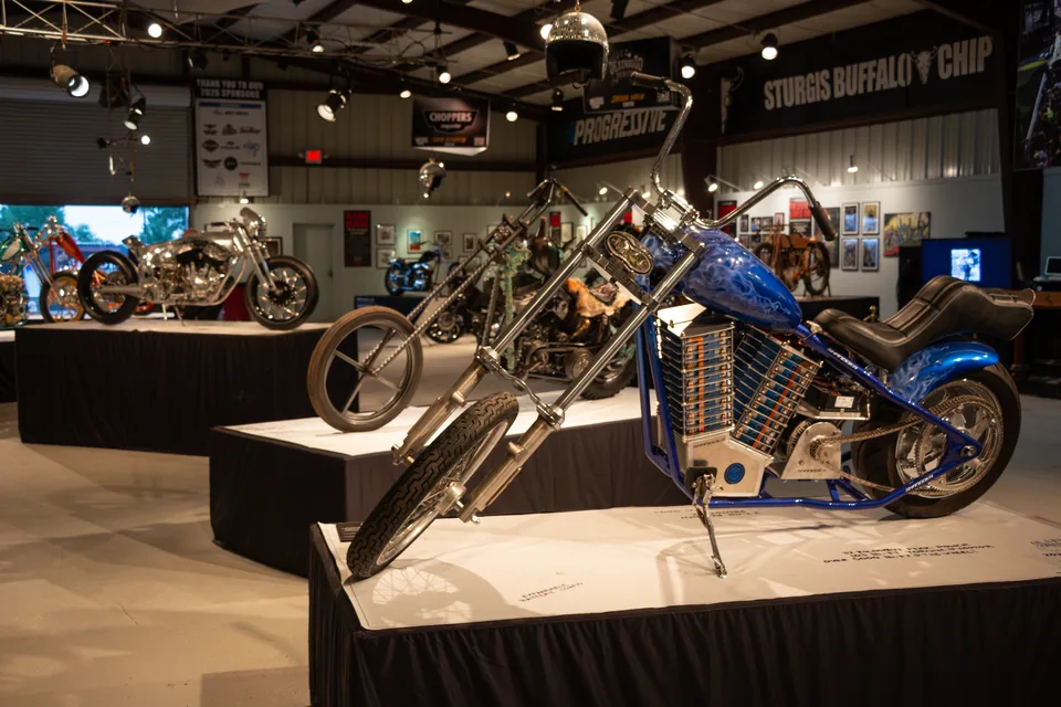 El chopper sobre su soporte en la exposición, con el salón y otras motos alrededor.