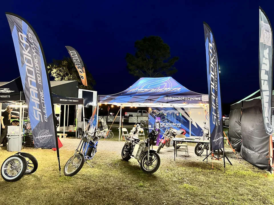 El stand de Shandoka en un evento de noche, con banderas de la marca a cada lado y motos estacionadas al frente.