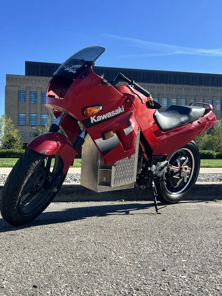 La Ninja 250 eléctrica en tres cuartos frontal y desde abajo: parabrisas, carenado rojo y, debajo, baterías y motor de buje.