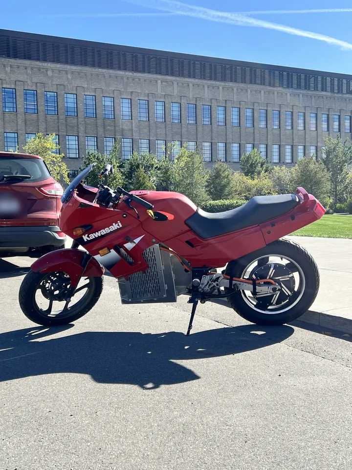 The electric Kawasaki Ninja 250 parked on the street in its red bodywork, side profile, the vented battery pack below the fairing and the hub motor in the rear wheel.